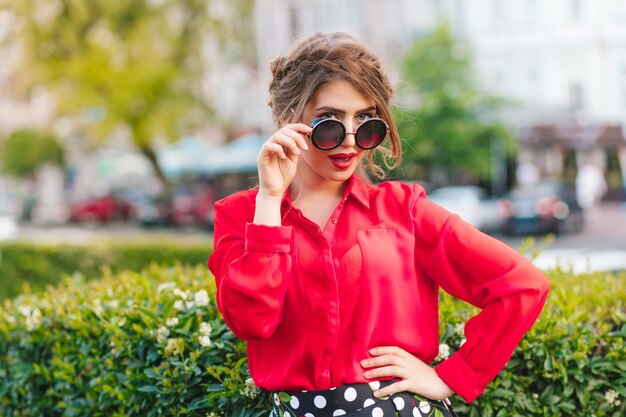 Retrato de niña bonita con gafas de sol posando para la cámara en el parque. Ella tiene peinado, blusa roja.