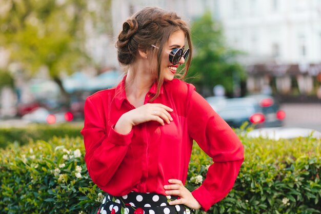 Retrato de niña bonita con gafas de sol posando para la cámara en el parque. Lleva blusa roja y un bonito peinado. Ella está sonriendo a un lado.