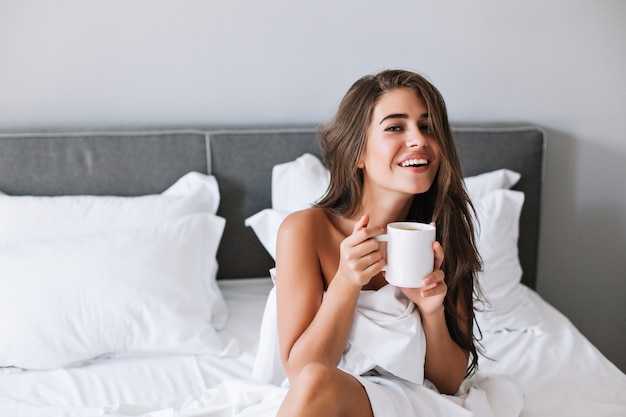 Retrato de niña bonita con hombros traviesos tomando café en la cama por la mañana en el apartamento.