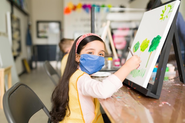 Retrato de una niña bonita con una máscara facial que está sentada con un pincel frente a una pintura que está haciendo en su escuela de arte