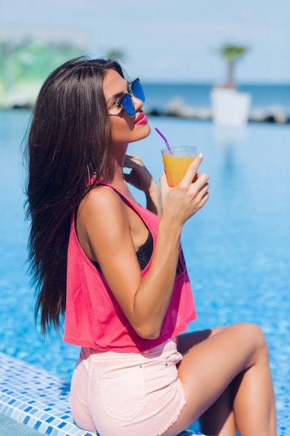 Retrato de niña bonita morena con pelo largo está sentado junto a la piscina. Ella sostiene la bebida y mantiene los ojos cerrados.