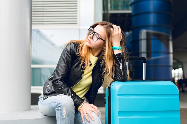 Foto gratuita retrato de niña bonita con pelo largo con gafas sentado afuera en el aeropuerto. viste un suéter amarillo con chaqueta negra y jeans. se inclinó hacia la maleta y mira a lo lejos.