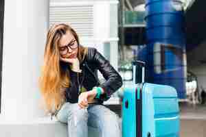 Foto gratuita retrato de niña bonita con pelo largo con gafas sentado afuera en el aeropuerto. viste un suéter amarillo con chaqueta negra y jeans. se inclinó hacia la maleta y mira el reloj aburrida.