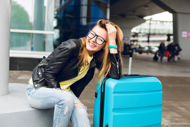 Retrato de niña bonita con pelo largo con gafas sentado afuera en el aeropuerto. Viste un suéter amarillo con chaqueta negra y jeans. Se inclinó hacia la maleta y sonrió a la cámara.