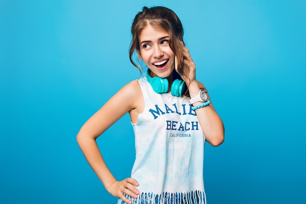 Retrato de niña bonita con pelo largo y rizado en la cola hablando por teléfono sobre fondo azul en estudio. Lleva camiseta blanca y auriculares azules en los hombros.