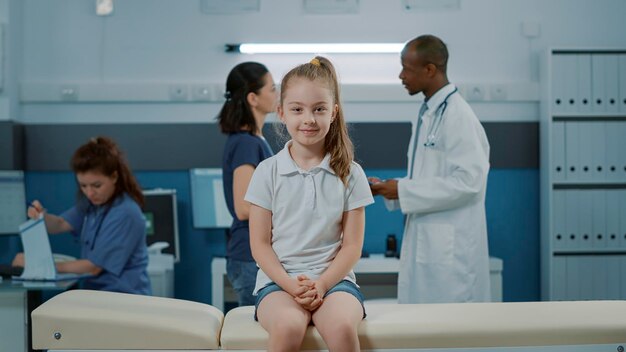 Retrato de una niña en la cama esperando para comenzar el examen de chequeo en el gabinete médico con el médico. Niño pequeño con enfermedad mirando la cámara y preparándose para una cita de visita de control en el consultorio.