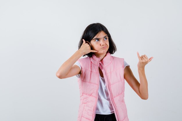 Retrato de una niña en camiseta blanca y chaleco acolchado mostrando gesto de teléfono