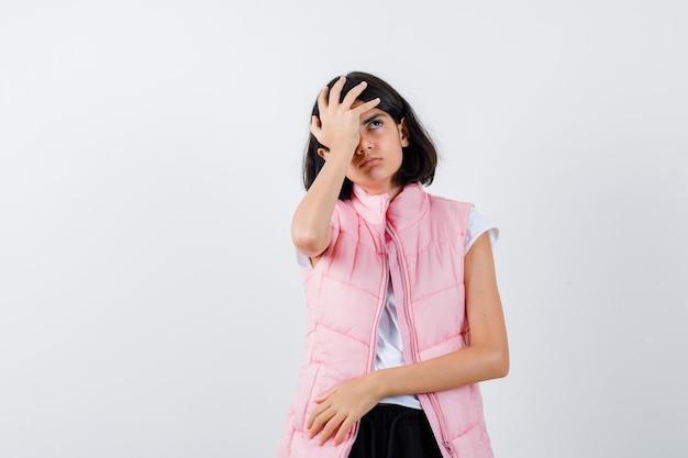 Retrato de una niña en camiseta blanca y chaleco acolchado poniendo la mano en la cabeza.