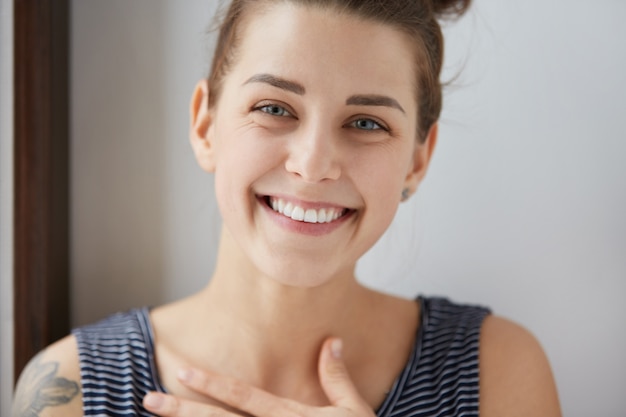 Retrato de niña caucásica feliz con moño de cabello morena, tatuaje en el hombro y top despojado. Mujer atractiva sonriendo con todos sus dientes blancos, poniendo su mano sobre el pecho con alegría sin fin.