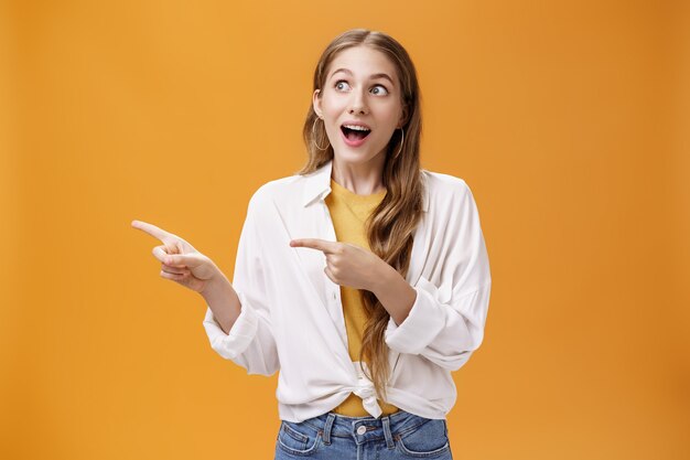 Retrato de niña caucásica guapa divertida y emocionada con peinado ondulado justo en blusa sobre camiseta mirando y apuntando a la izquierda asombrada y sorprendida posando sobre fondo naranja.