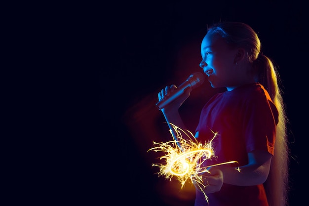 Retrato de niña caucásica sobre fondo oscuro de estudio en luz de neón. Hermoso modelo femenino con altavoz y bengala.