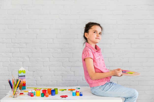 Retrato de una niña contemplada sentada en la mesa blanca mezclando la acuarela en la paleta