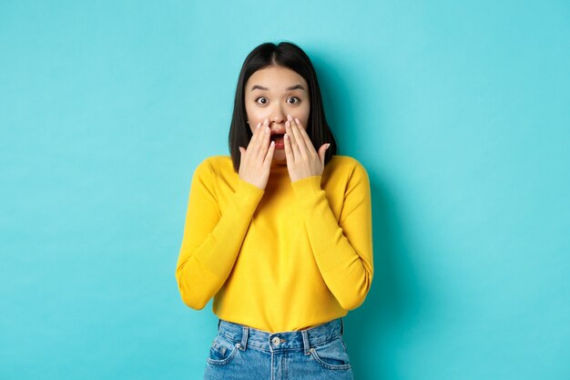 Retrato de niña coreana impresionado diciendo wow, jadeando y mirando asombrado a la cámara, de pie en jersey amarillo sobre fondo azul.