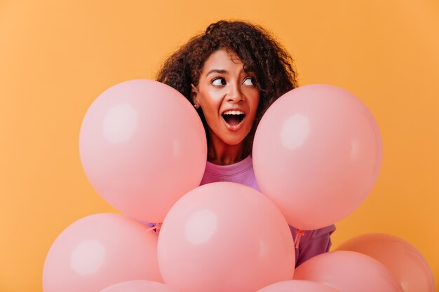 Retrato de niña de cumpleaños sorprendida mirando a otro lado mientras posa con globos. Dama africana divertida jugando durante la fiesta.