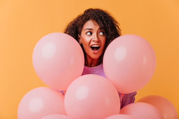 Retrato de niña de cumpleaños sorprendida mirando a otro lado mientras posa con globos. Dama africana divertida jugando durante la fiesta.