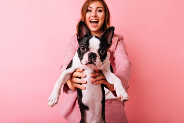 Retrato de niña despreocupada con estilo con perrito gracioso en primer plano. Encantadora dama caucásica con cabello oscuro que expresa buenas emociones durante la sesión de retratos con bulldog francés.