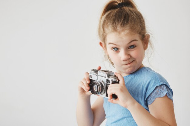 Retrato de niña divertida con cabello rubio en peinado de cola, con expresión tonta, sosteniendo la cámara en las manos va a tomar una foto.