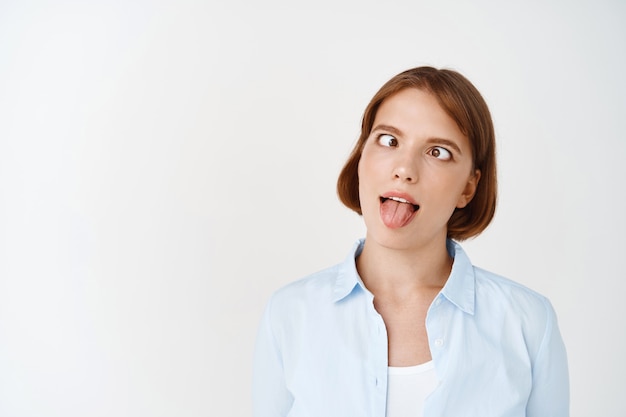 Retrato de niña divertida y juguetona entrecerrando los ojos y mostrando la lengua tonta, haciendo muecas, vistiendo una blusa de oficina azul, teniendo una emoción loca, de pie contra la pared blanca