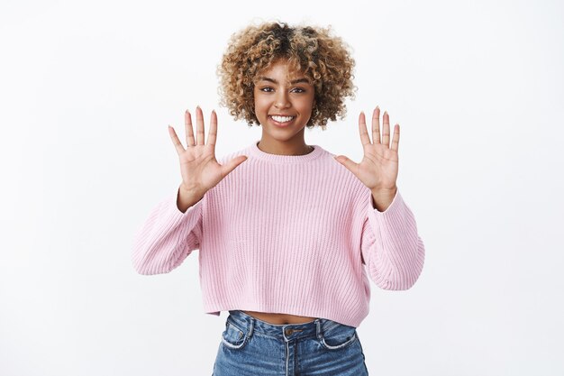 Retrato de niña elegante afroamericana optimista y alegre con corte de pelo afro rubio sonriendo encantada y segura mientras levanta las manos y muestra el número diez, posando contra la pared blanca