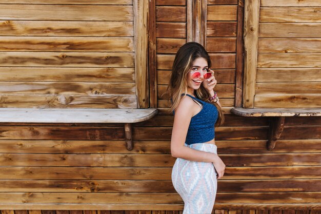 Retrato de niña elegante bien construida en falda midi y gafas de sol coloridas sonriendo, de pie con expresión de sorpresa