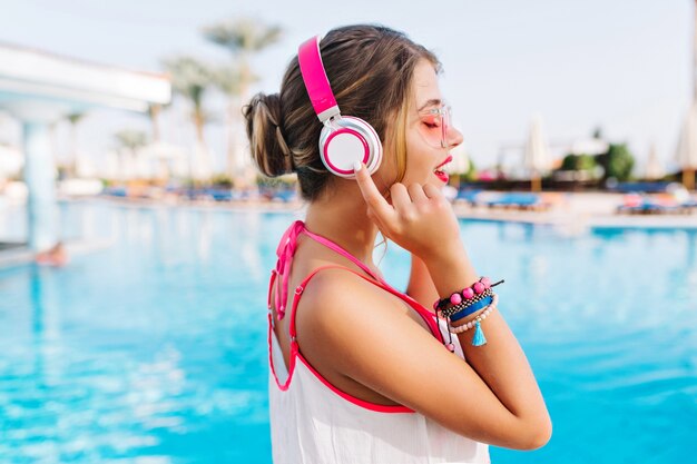 Retrato de niña elegante en camiseta blanca pasar tiempo cerca de la piscina al aire libre disfrutando de música y aire fresco