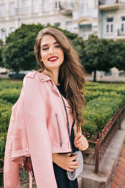 Retrato de niña elegante con pelo largo y rizado posando. Lleva vestido negro, chaqueta rosa, bolso de mano, labios rojos. Ella esta sonriendo .