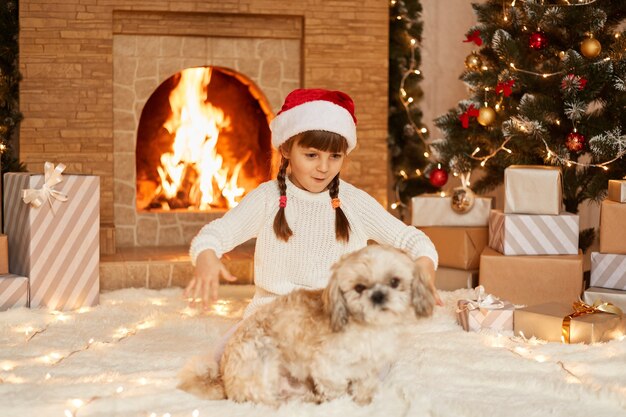 Retrato de niña emocionada asombrada con suéter blanco y sombrero de santa claus, jugando con su cachorro en la sala festiva con chimenea y árbol de Navidad.