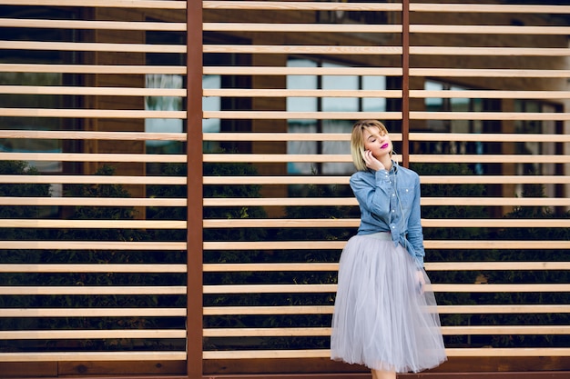 Retrato de una niña de ensueño con el pelo rubio corto volador y labios rosados brillantes escuchando música en un teléfono inteligente con balks de madera a rayas detrás