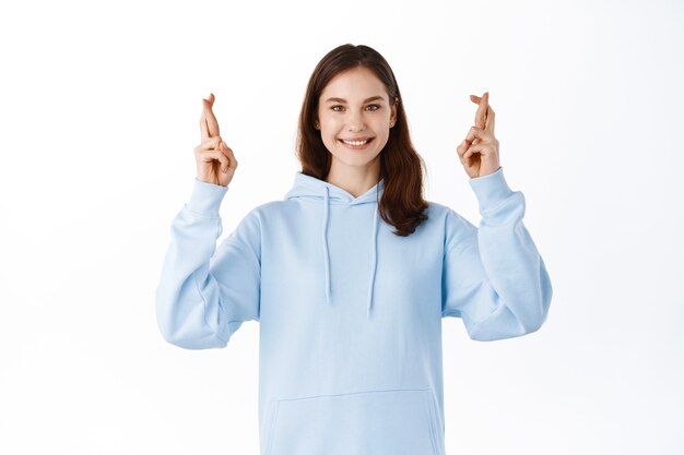 Retrato de niña esperanzada que desea aprobar el examen, cruzar los dedos, buena suerte y mirar con esperanza y sonrisa positiva al frente, sueño hecho realidad, de pie contra la pared blanca