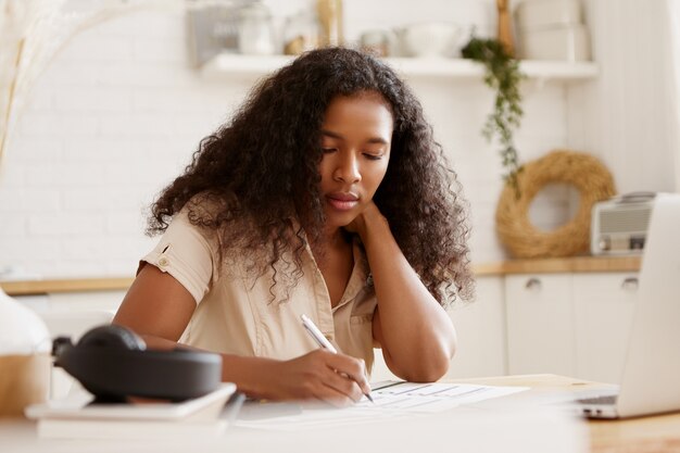 Retrato de niña estudiante afroamericana seria concentrada sosteniendo un lápiz, escribiendo, preparándose para los exámenes o haciendo la tarea en la cocina, sentada en la mesa del comedor con una computadora portátil abierta y libros