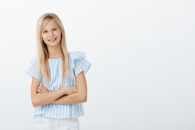 Foto gratuita retrato de niña europea adorable confiada en blusa azul, tomados de las manos cruzadas sobre el pecho y sonriendo con expresión segura de sí misma, siendo enérgica y alegre