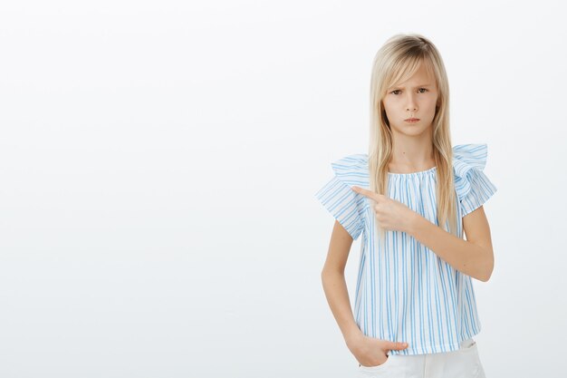 Retrato de niña europea enojada ofendida con cabello rubio, frunciendo el ceño y mirando por debajo de la frente, apuntando a la esquina superior izquierda mientras está de pie sobre la pared gris molesta