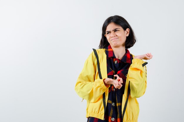 Retrato de niña extendiendo las palmas en camisa a cuadros, chaqueta y mirando insatisfecho vista frontal