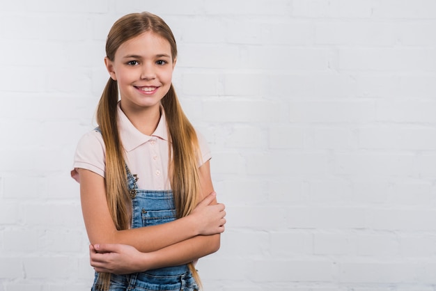 Retrato de una niña feliz con el brazo cruzado mirando a la cámara de pie contra la pared blanca