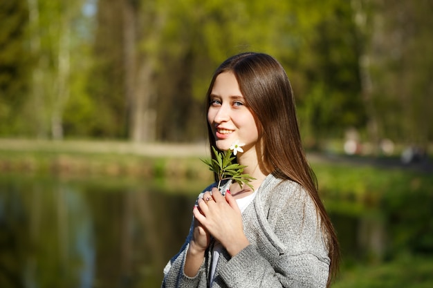 Retrato de una niña feliz en el fondo de un parque con un lago, sosteniendo una flor de bosque salvaje. Primavera día soleado