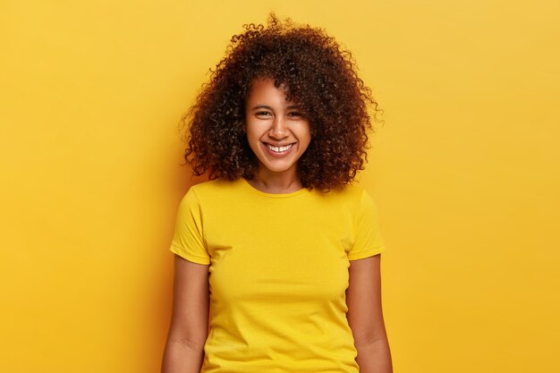 Retrato de niña feliz hipster ríe positivamente, se ríe y sonríe despreocupadamente, viste ropa casual de color amarillo brillante, tiene poco espacio entre los dientes, se siente encantada, posa en interiores, tiene belleza natural