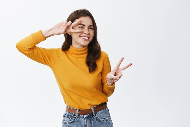 Retrato de niña feliz posando en estudio con signo v, mostrando gesto de paz cerca de los ojos y sonriendo despreocupado, disfrutando de la vida, de pie alegre contra la pared blanca