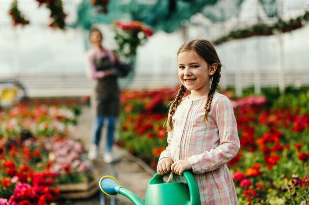 Retrato de niña feliz con regadera en un invernadero La madre está trabajando en segundo plano