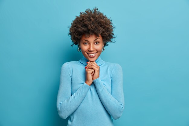 Retrato de niña feliz tiene cabello afro mantiene las manos debajo de la barbilla se ve con expresión satisfecha y complacida