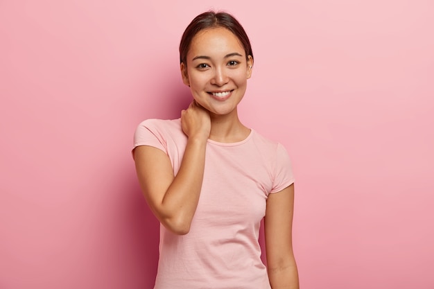 Retrato de niña femenina con sonrisa agradable, mirada gentil, toca el cuello, tiene expresión de la cara encantada, tiene una conversación agradable con un amigo cercano, vestida con ropa informal, aislada en la pared rosa