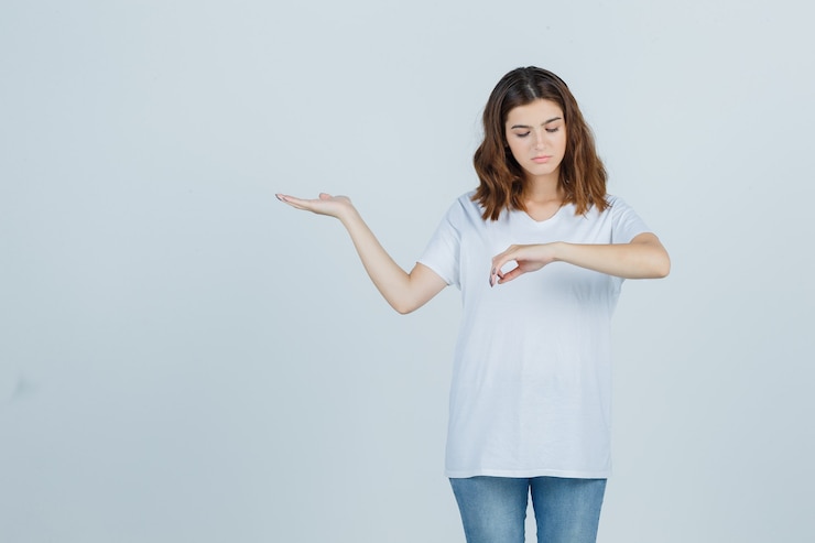 Foto gratuito retrato de niña fingiendo mirar su reloj, extendiendo la palma hacia un lado en camiseta blanca y mirando pensativa vista frontal