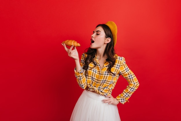 Retrato de niña francesa rizada comiendo croissant crujiente en la pared roja. Mujer de cabello oscuro con blusa a cuadros y sombrero amarillo mira moño.