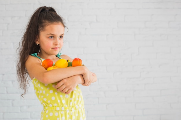 Retrato de niña con frutas de pie contra la pared de ladrillo blanco