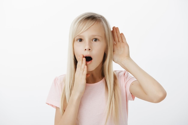 Retrato de niña habladora intrigada sorprendida con cabello largo y rubio, sosteniendo la mano cerca de la oreja y boca abierta, diciendo wow mientras escucha una conversación, oye rumores o chismes de un amigo