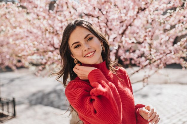 Retrato de niña hermosa en suéter rojo cerca de sakura. Encantadora mujer en traje de cashemere sonriendo y mirando a puerta cerrada en el jardín