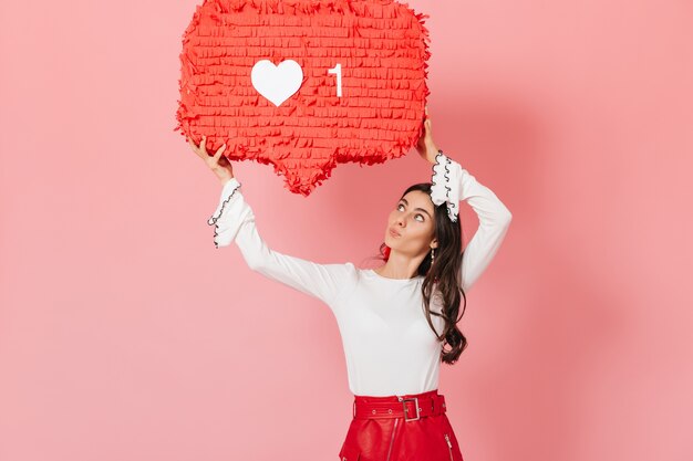 Retrato de niña con interés mirando enorme letrero "" como "" de Instagram. Morena en falda roja posando sobre fondo rosa.