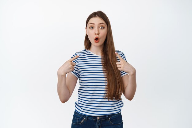 Retrato de niña jadeando con asombro, sorprendida y señalándose a sí misma, sorprendida por algo que le sucedió, de pie sobre fondo blanco.