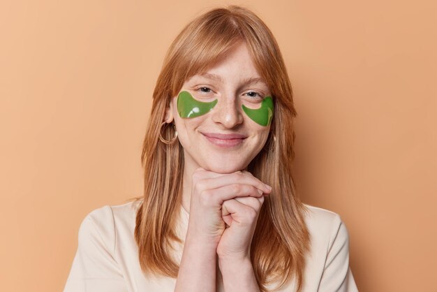 Retrato de niña de jengibre satisfecha sonríe suavemente mantiene las manos debajo de la barbilla aplica parches de hidrogel verde debajo de los ojos se somete a tratamientos de cuidado de la piel vestidos casualmente aislados sobre fondo marrón