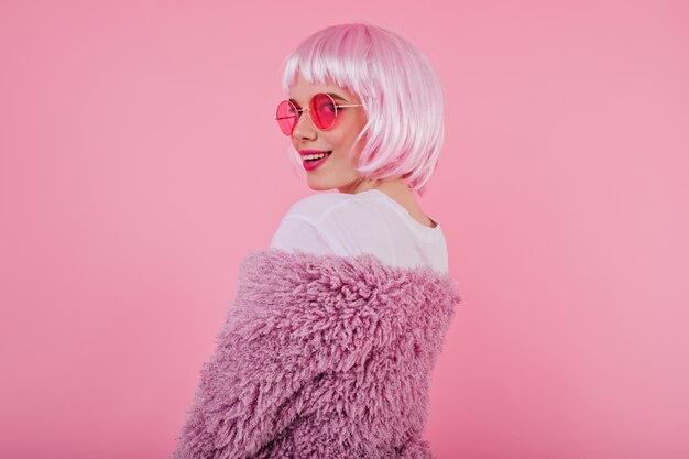 Retrato de niña juguetona en peruke rosa brillante y gafas de sol. Debonair mujer caucásica mirando por encima del hombro con una sonrisa