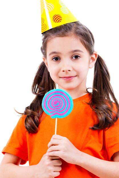 Retrato de niña linda en camiseta naranja y gorro de fiesta con caramelo de colores - aislado en blanco